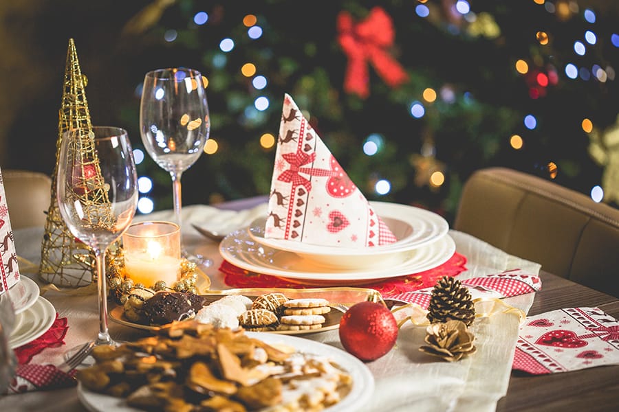 A festive holiday table set with plates, folded decorative napkins, wine glasses, cookies, candles, and ornaments, with a Christmas tree glowing in the background