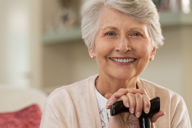 An elderly woman with short gray hair smiles warmly while resting her hands on the handle of a cane