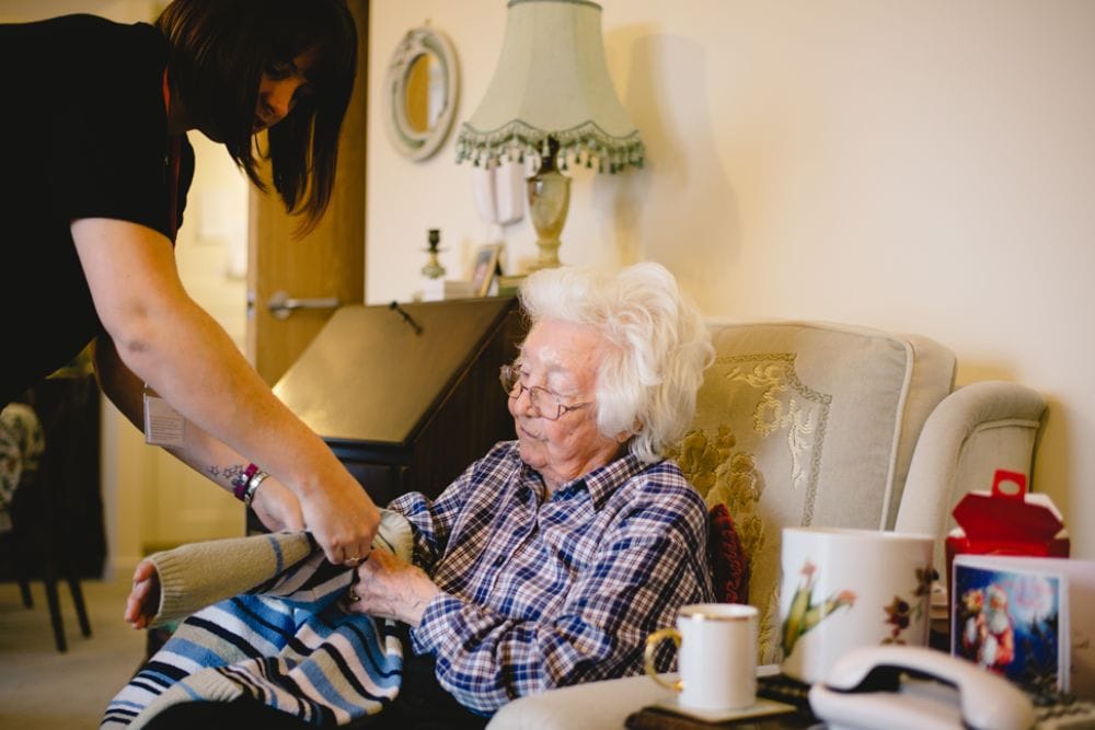 Live-In-Care-Colleague An elderly lady sits in an armchair. A Unique Senior Care colleague helps the client to put on a jumper.