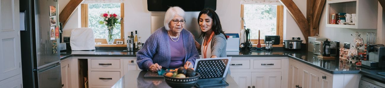 kitchen-laptop-1300x300 A senior lady stands at a grey kitchen island with a laptop. Beside her, a dark haired caregiver with an orange lanyard around her neck.