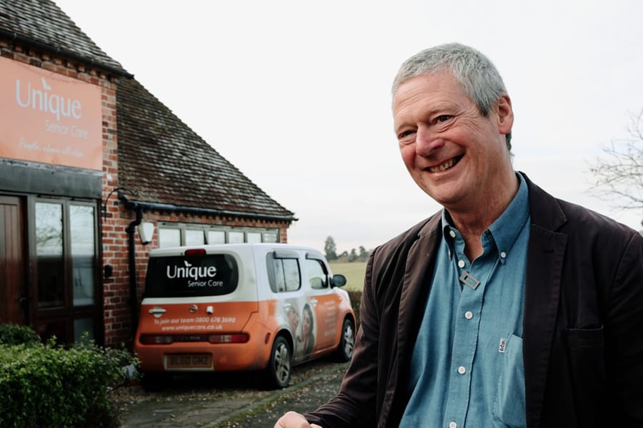 A smiling man stands outside a Unique Senior Care building, with a branded company vehicle parked nearby.