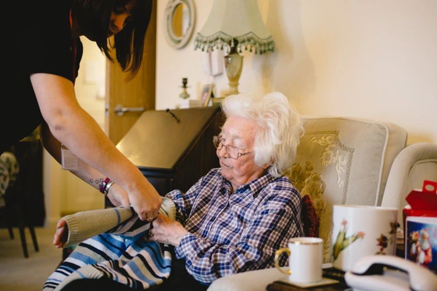 A caregiver with dark hair helps an elderly woman with white hair put on a knitted cardigan in her cosy living room in Somerset. The room features a floral lampshade and a side table with a mug, illustrating the importance of ensuring compatibility in live-in care.