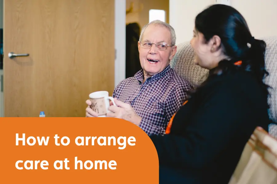 A smiling elderly man holding a mug sits on a sofa, chatting with a caregiver wearing a black uniform with an orange lanyard. The image has an orange text overlay that reads, "How to arrange care at home."