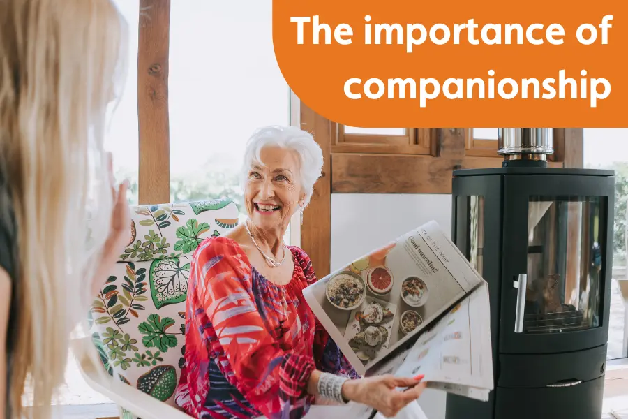 An older woman sat smiling reading a newspaper, while showing her Caregiver, overlayed with the text 'the importance of companionship'