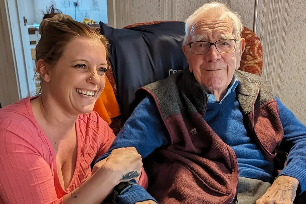 A young caregiver sits beside an elderly man in glasses, both smiling as they enjoy a friendly moment together.