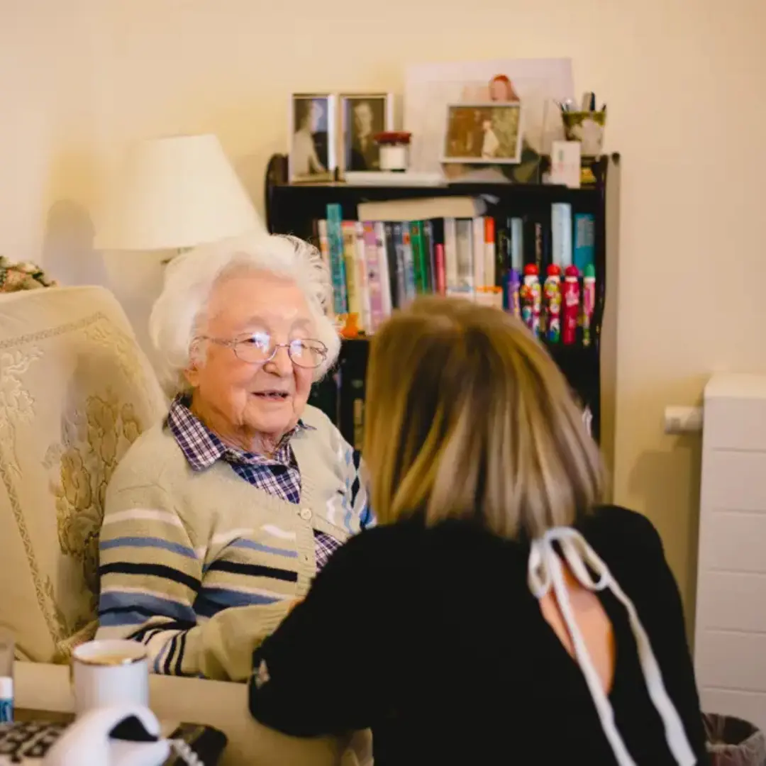 An elderly lady sat smiling in her chair while a caregiver kneels down to talk to her