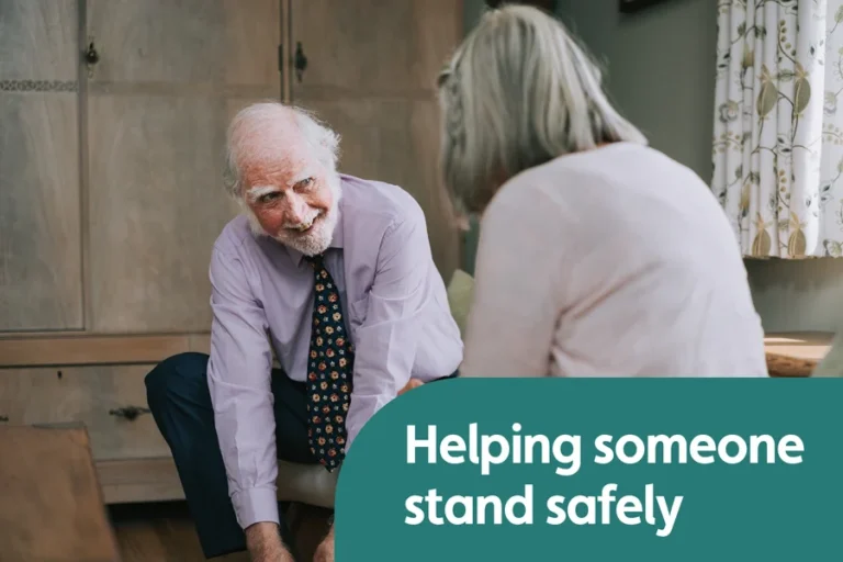Older man seated at home speaking with another person before getting up from a chair.