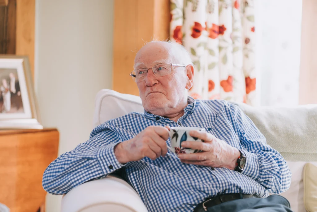 An elderly man sat on a sofa with a cup of tea, with a thoughtful expression