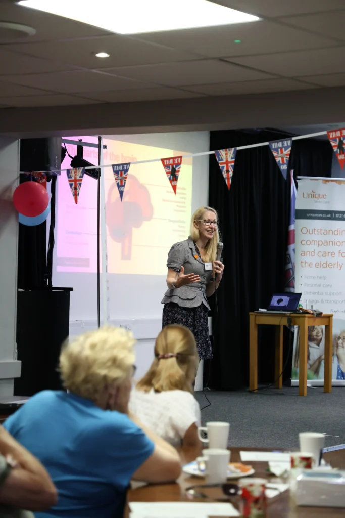 Jo Cleary, Learning and Development Manager at Unique Senior Care, delivering a presentation on dementia during the support event in Knowle. She stands at the front of the room with a microphone, gesturing as she speaks, while attendees listen at tables. Union Jack bunting and a projection screen are visible in the background.