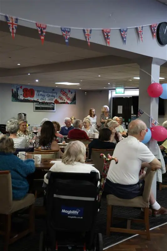 Local residents gathered at the Dementia Awareness and Support Event in Knowle, seated around tables listening to a speaker. The room is decorated with red and blue balloons and Union Jack bunting, creating a friendly, community-focused setting.