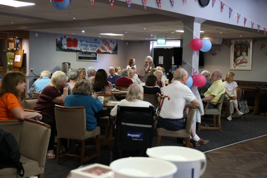 Attendees seated around tables at the Dementia Awareness and Support Event in Knowle, engaging in conversation and listening to a speaker. The room is decorated with bunting and balloons, creating a warm and welcoming atmosphere.