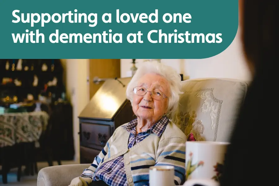Older woman sitting in an armchair, smiling while talking with a family member in a Christmas decorated living room.