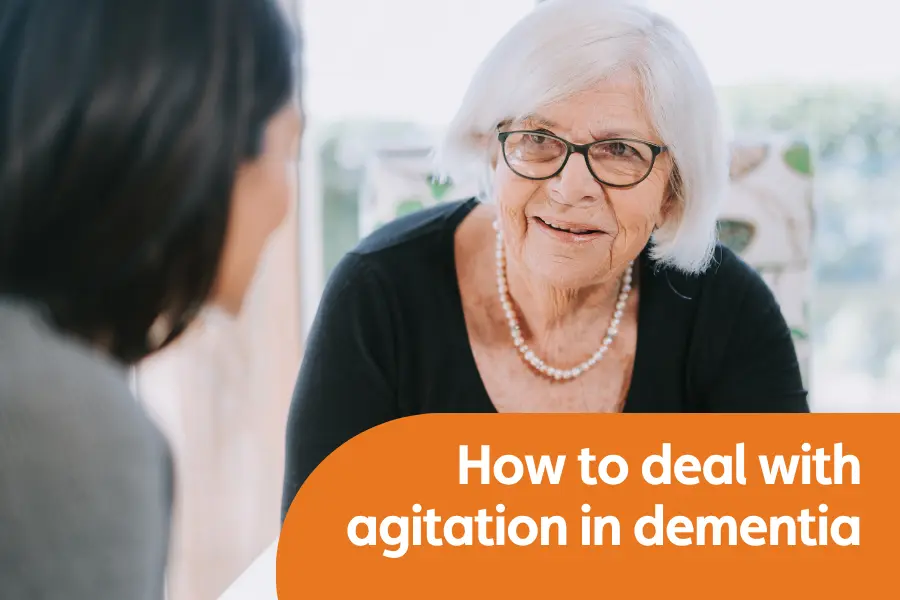 Featured image for blog on how to deal with dementia agitation. Image shows an older woman with white hair smiling at her caregiver who is out of focus and off to the side