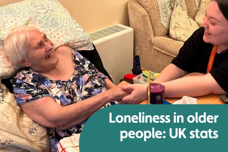 Featured for image for blog on loneliness stats in the UK. Image shows elderly lady in bed holding hands with her carer . theyre both smiling at each other warmly.