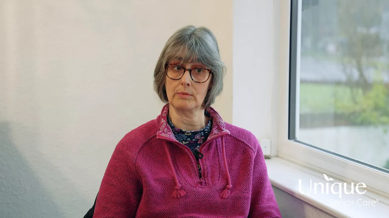 Woman in a pink sweater explaining how consistent, longer home-care visits improved wellbeing, seated by a window