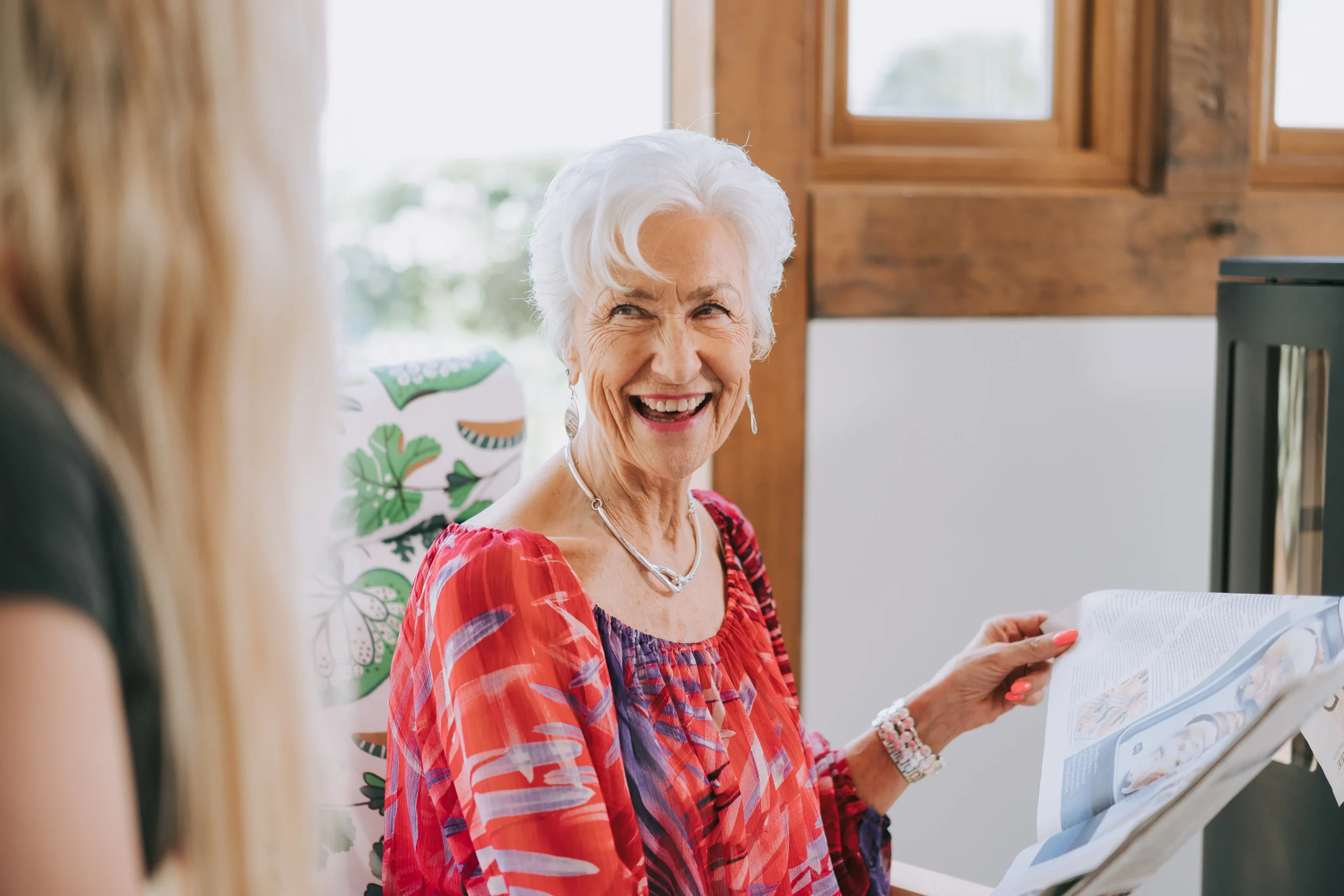 An older lady with white hair smiling while holding an open newspaper. Shes looking towards someone who is just out of shot with their back to the camera.