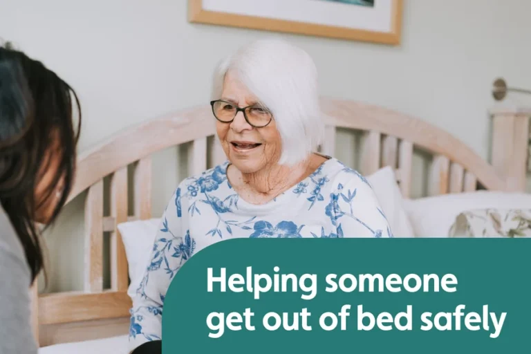 Older woman sitting upright on a bed while a Caregiver talks with her nearby.