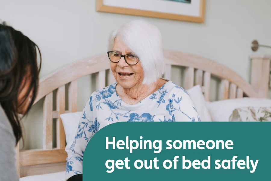 Older woman sitting upright on a bed while a Caregiver talks with her nearby.