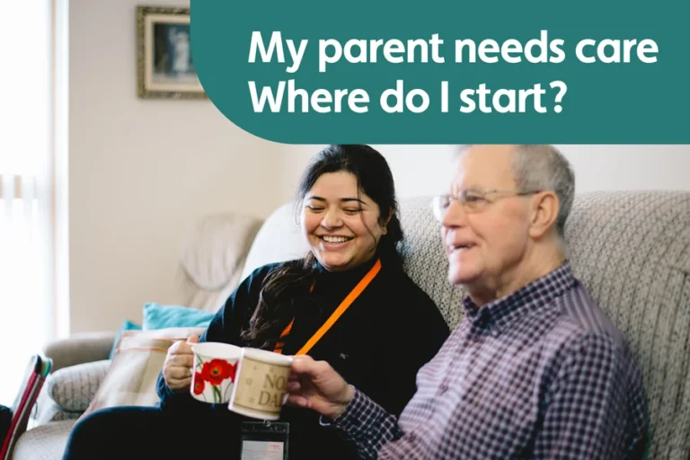 Caregiver and older man sitting together at home, smiling over cups of tea.