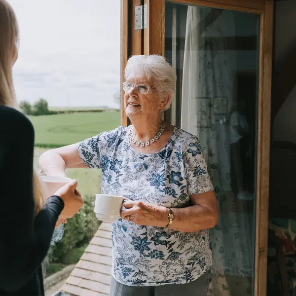An older woman stood by a window smiling at her caregiver while holding a cup of tea
